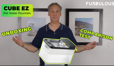 A man stands indoors holding a white and black Cube Ez pet water fountain, with graphic overlays reading 'UNBOXING', 'COMPARISON TEST', and 'CUBE EZ Pet Water Fountain'.